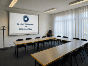 Ein moderner, unbesetzter Konferenzraum, dessen Tische in U-Form angeordnet sind. Die Projektionsfolie zeigt den Willkommenstext: „Herzlich Willkommen zur KI-Weiterbildung“ und das KI-Weiterbildungszentrum-Logo.